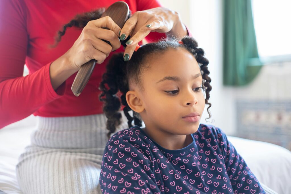 A mother lovingly styles her daughter's hair, creating braided pigtails in a warm indoor setting.